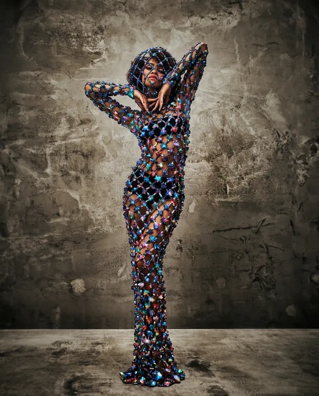 A striking editorial photograph features a model in a shimmering, sheer gown adorned with colorful gemstones, posing elegantly against a textured gray wall. The composition evokes a glamorous and artistic mood, with vibrant hues contrasting against the muted background.