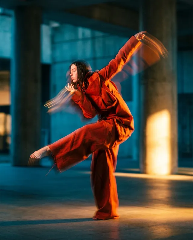 This editorial photograph captures a dancer in mid-motion, clad in an orange outfit, set against a backdrop of concrete columns. The dynamic pose conveys a sense of energy and grace, enhanced by warm, golden lighting that creates a captivating contrast with the cool tones of the environment.