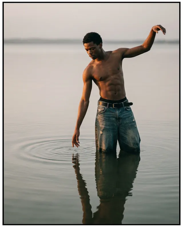 A contemplative portrait of a man standing in shallow water, creating ripples as he interacts with the surface. The mood is serene with soft, natural tones dominating the image.