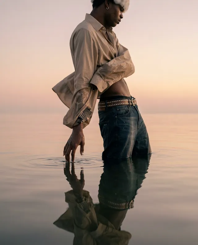 A fashion photograph features a male model standing in shallow water at dusk, his reflection visible in the calm surface. The soft pastel colors of the sunset create a serene mood, highlighting the model's stylish attire and dynamic pose.