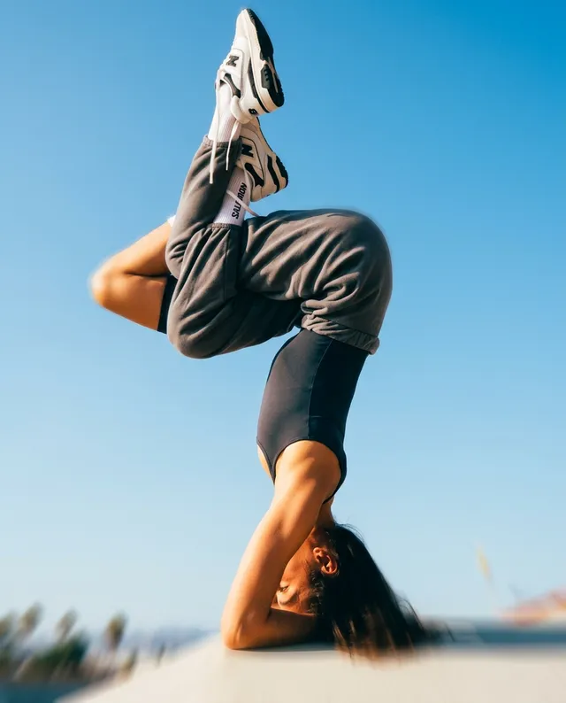 A dynamic editorial photograph captures a woman performing a headstand against a clear blue sky. The image exudes an energetic mood, featuring vibrant colors with a focus on the cool tones of the sky and the neutral greys of her outfit.