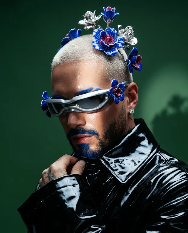 A striking editorial photograph features a person with a stylish, platinum buzz cut and bold blue facial hair, adorned with floral accessories and wearing futuristic sunglasses. The mood is eclectic and avant-garde, dominated by deep greens, blues, and metallic highlights.