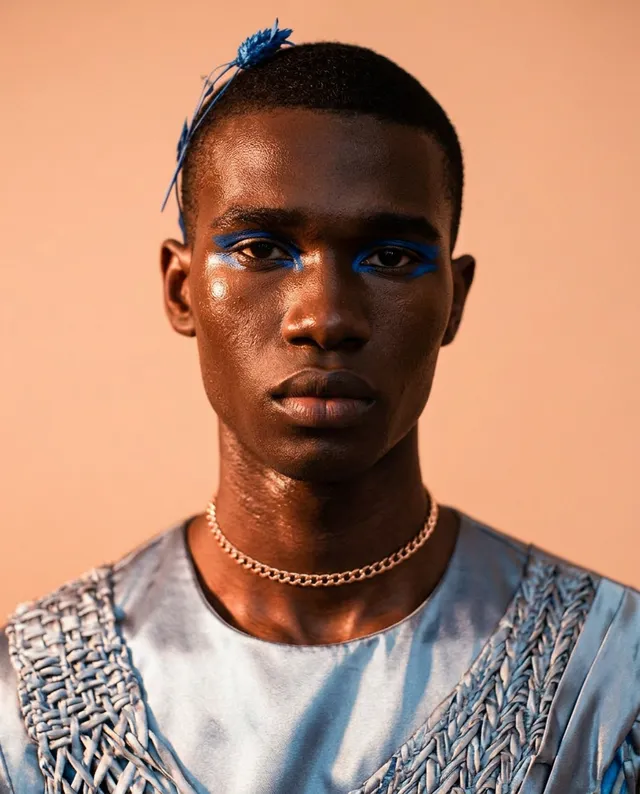 A striking editorial portrait featuring a young man with bold blue eyeshadow and a delicate accessory in his hair, set against a soft, warm background. The mood is confident and artistic, with dominant shades of blue and earth tones.