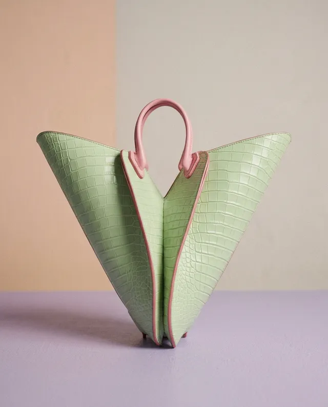 A stylish, pastel green handbag with a unique shape and pink accents sits against a softly colored background, evoking a chic and playful mood. The image features clean lines and a minimalist aesthetic, highlighting the bag's texture and design.