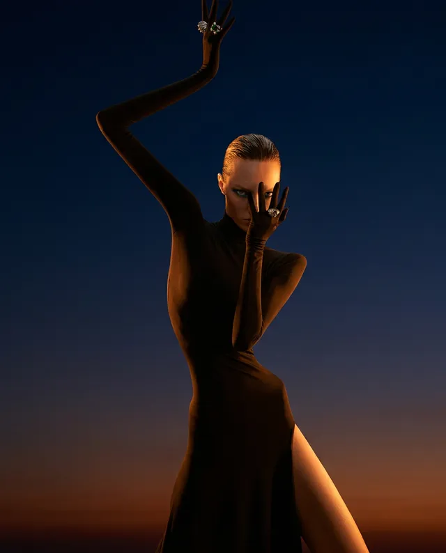 A striking editorial photograph featuring a model in a sleek black gown, accentuated by dramatic poses and bold jewelry against a gradient sunset backdrop. The mood is alluring and mysterious, with deep blues and warm oranges dominating the scene.