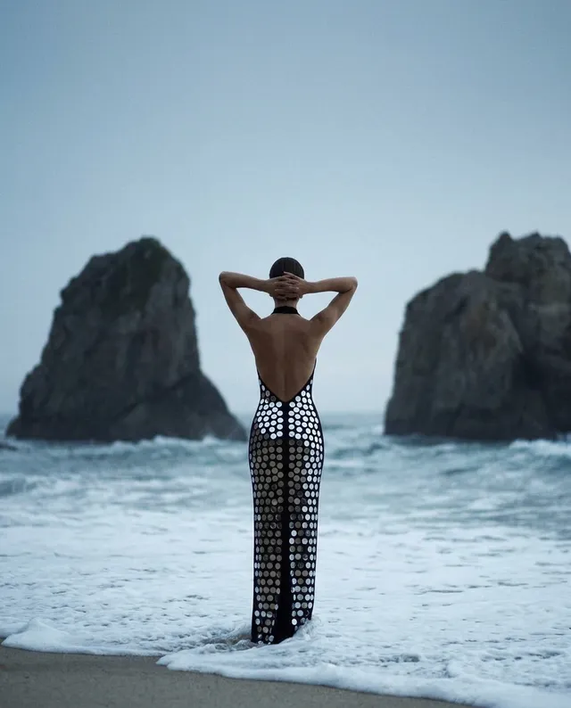A stunning editorial photograph captures a model standing at the ocean's edge in a striking backless dress adorned with reflective discs, exuding a serene yet bold mood. The cool, muted tones of blue and gray contrast with the shimmering silver of the dress, creating a captivating visual ambiance.