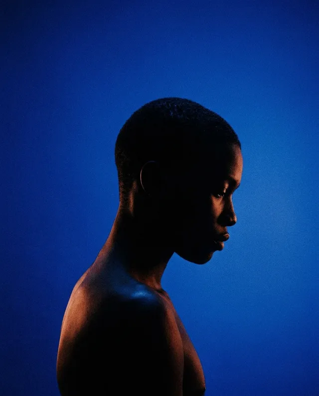 A striking editorial photograph of a young person in profile, illuminated against a deep blue background. The mood is contemplative, with contrasting warm tones highlighting the subject's silhouette.