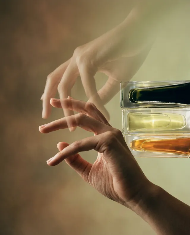 A close-up of a delicate hand reaching towards stacked glass vials, capturing an ethereal and minimalist mood. The image features soft, muted tones with earthy greens and warm amber highlights, blending editorial photography with a painterly quality.