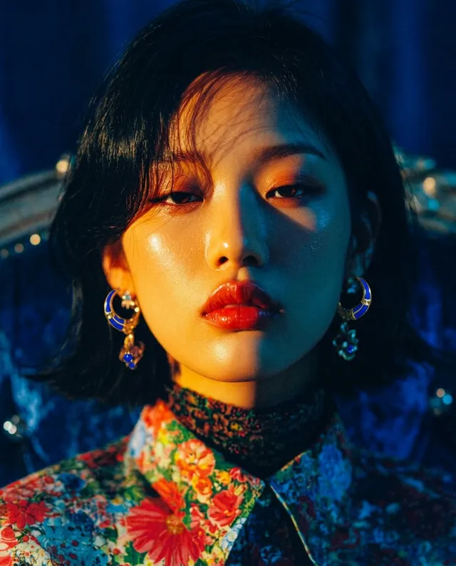 A portrait of a young woman with striking features, styled in a vibrant floral blouse and bold accessories, exuding a confident and intense mood. The image is characterized by rich, saturated colors with a play of warm and cool lighting, enhancing the dramatic atmosphere.