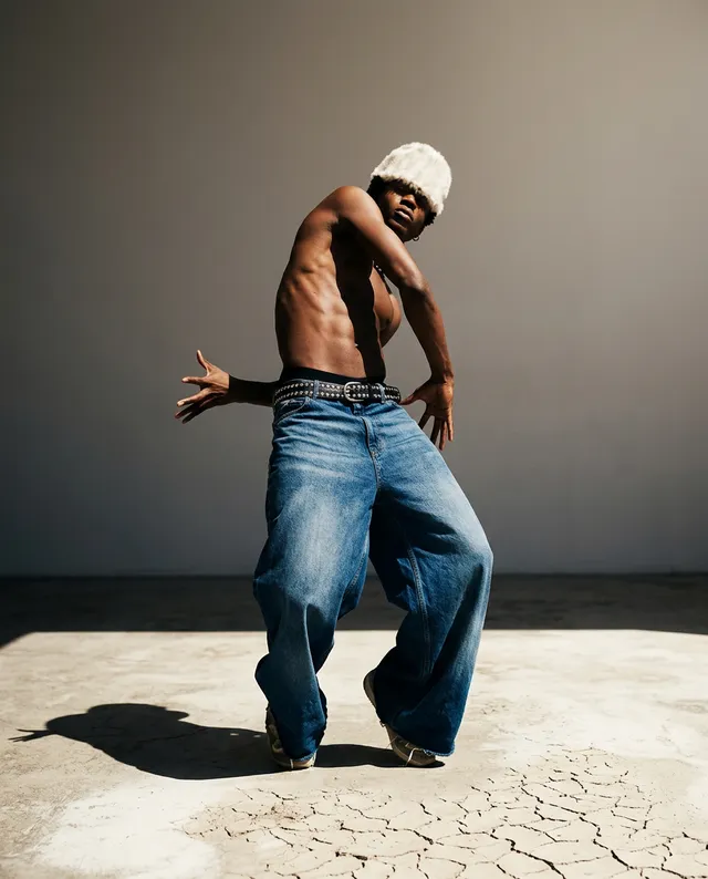 A dynamic editorial photograph features a bare-chested dancer in oversized denim pants, showcasing fluid movement against a minimalist, sandy backdrop. The image conveys a bold and energetic mood, highlighted by earthy tones and stark contrasts.