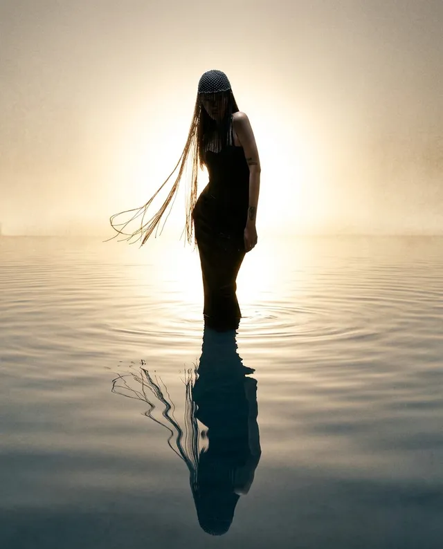An ethereal editorial photograph features a silhouetted figure in a flowing black dress, standing in tranquil water against a glowing backdrop. The serene mood is emphasized by soft, muted tones and the interplay of light and reflection.