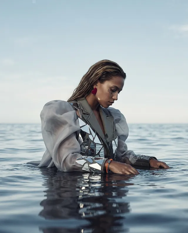 A reflective editorial photograph captures a woman wading in calm water, dressed in a striking outfit with metallic accents and exaggerated sleeves. The mood is serene and introspective, dominated by soft blues and neutrals.