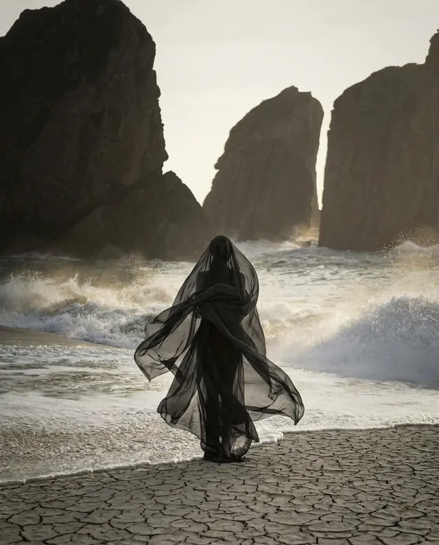 A mysterious figure in a flowing black veil stands at the shoreline, surrounded by dramatic rock formations and crashing waves. The image, captured in a moody, ethereal style, features a palette of dark tones contrasted with the soft light of the setting sun.