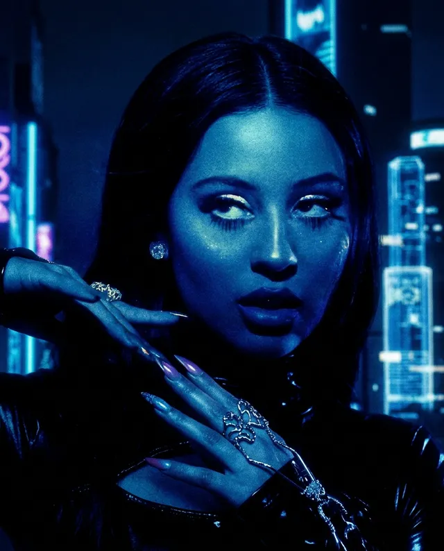 A striking editorial photograph featuring a woman with bright blue-toned lighting, exuding an edgy and futuristic vibe. The image highlights her dramatic expression and metallic accessories against a backdrop of neon city lights, creating a moody atmosphere dominated by deep blues and blacks.