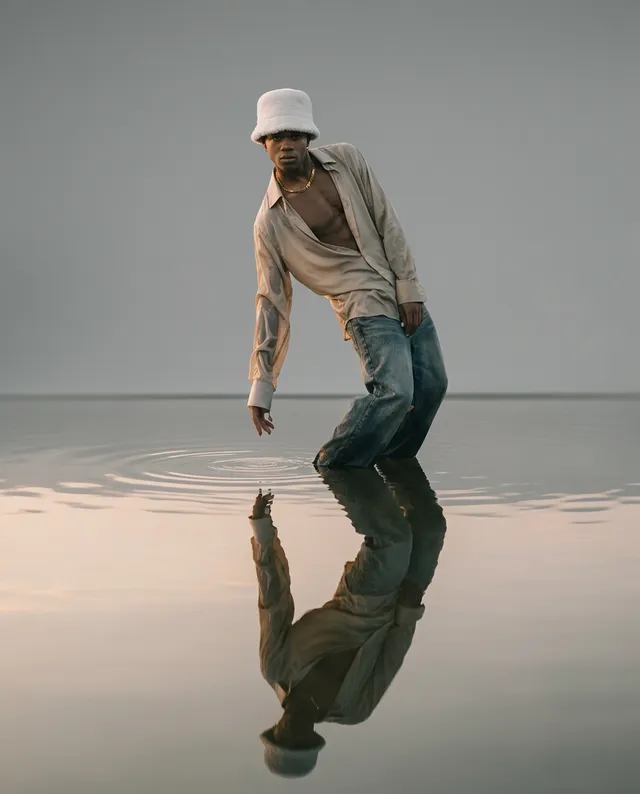 A lone figure in a loose, light-colored shirt and bucket hat stands against a serene, reflective surface, creating a tranquil yet contemplative mood. The image features soft hues of gray and beige, emphasizing a minimalist aesthetic and harmony with nature.