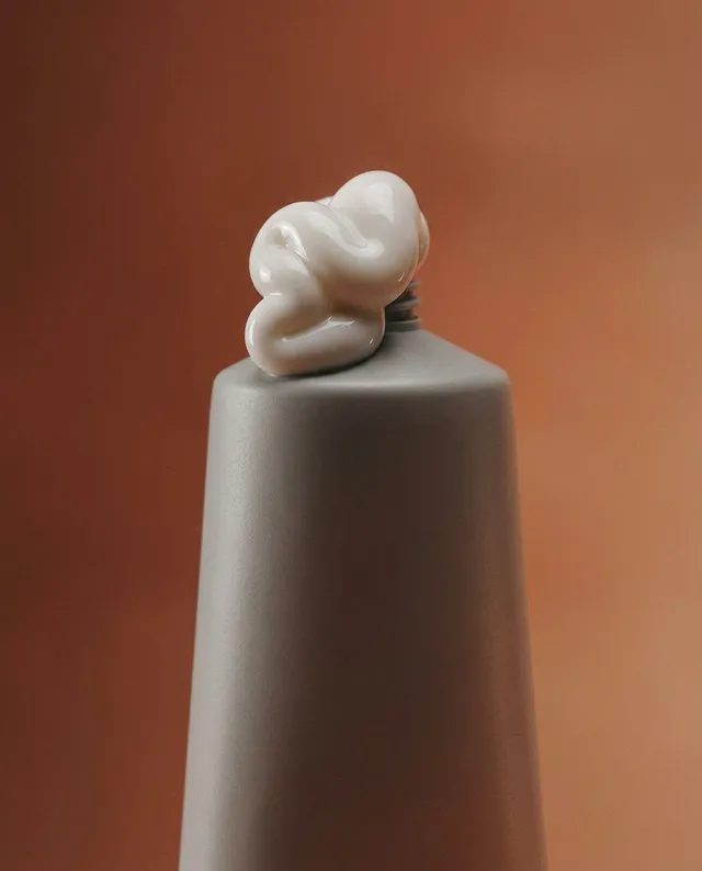 A minimalist still life featuring a gray ceramic vase topped with a smooth, white sculptural form, set against a warm, earthy background. The composition evokes a sense of calm and modern elegance through its soft lines and muted color palette.