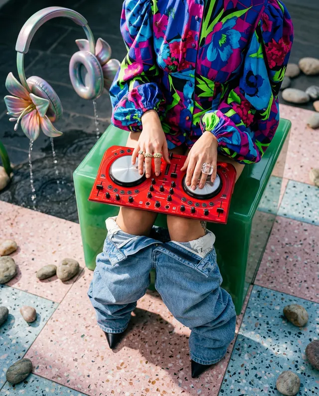 A vibrant editorial photograph showcases a person seated on a green block while mixing music on a striking red DJ controller, wearing a bold floral-patterned blouse and loose denim. The mood is playful and eclectic, dominated by bright colors like pink, green, and blue, accented by a whimsical setting featuring floral headphones and decorative stones.