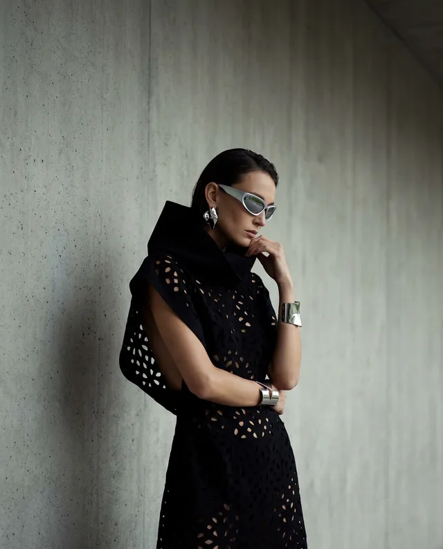 A poised model stands against a sleek, concrete wall, showcasing a striking black cut-out dress and metallic accessories. The editorial photography features a modern, fashion-forward aesthetic with a cool, minimalist atmosphere highlighted by deep blacks and subtle silver tones.