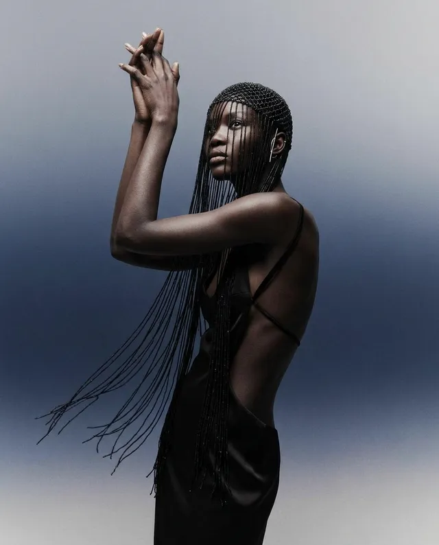 A striking editorial photograph featuring a model with intricately braided hair, posed gracefully against a gradient background that transitions from dark to light. The mood is both ethereal and bold, with dominant shades of black and soft pastels creating a dramatic contrast.