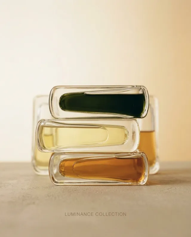 A minimalist editorial photograph featuring a stack of elegantly shaped glass bottles filled with vibrant oils in green, yellow, and amber hues. The soft, warm background enhances the serene and sophisticated mood of the Luminance Collection.
