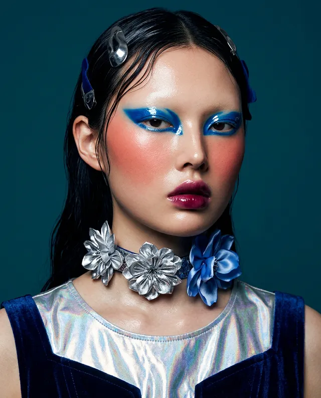 A striking editorial portrait featuring a model with bold blue eye makeup and bright pink cheeks, adorned with silver and blue floral accessories. The mood is edgy and vibrant, set against a deep teal background with dominant hues of blue, silver, and pink.