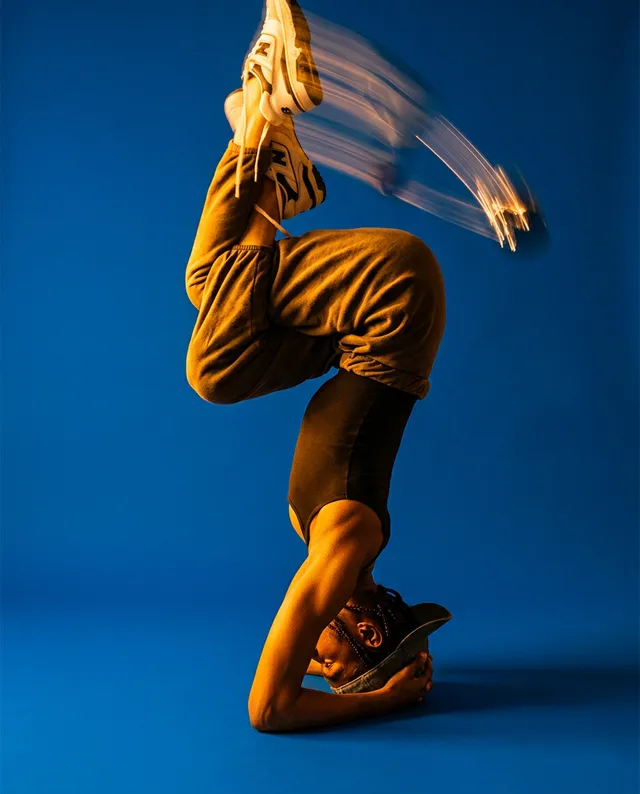 A dynamic editorial photograph captures a dancer in an impressive headstand against a vivid blue background, showcasing movement with a blur effect on the legs. The mood is energetic and vibrant, featuring contrasts of warm tones in the subject's attire against the cool backdrop.
