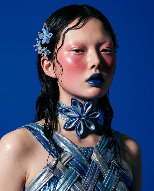 This editorial fashion photograph features a model with striking blue lips and exaggerated blush against a vivid blue backdrop, complemented by a floral hair accessory and intricate jewelry. The mood is bold and avant-garde, with a predominance of cool tones that enhance the high-fashion aesthetic.