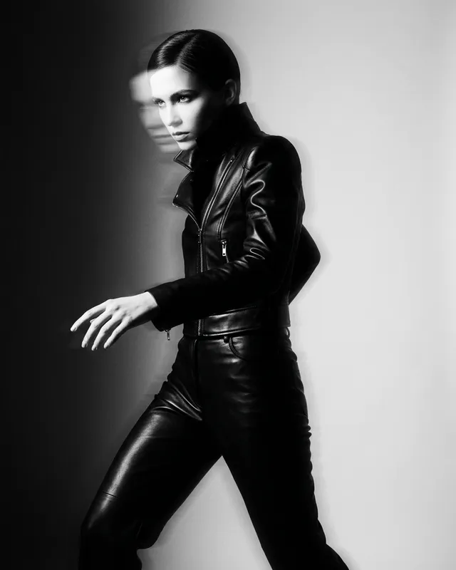 A striking editorial photograph features a model in a sleek black leather outfit, captured in a dynamic pose that conveys motion and intensity. The image employs high-contrast monochrome tones, enhancing the dramatic and edgy mood.