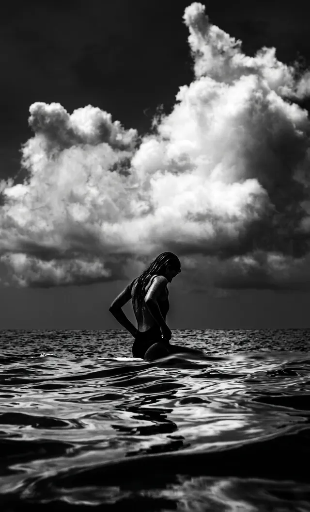 A striking black-and-white photograph captures a silhouetted figure wading through calm ocean waters, surrounded by dramatic clouds. The mood is contemplative, with contrasting tones of deep shadows and shimmering highlights on the water.