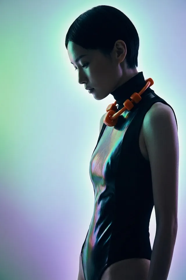 A stylish editorial photograph features a contemplative young woman in a sleek black bodysuit, complemented by an orange statement necklace. The mood is ethereal, with soft gradients of purple and green illuminating the background.