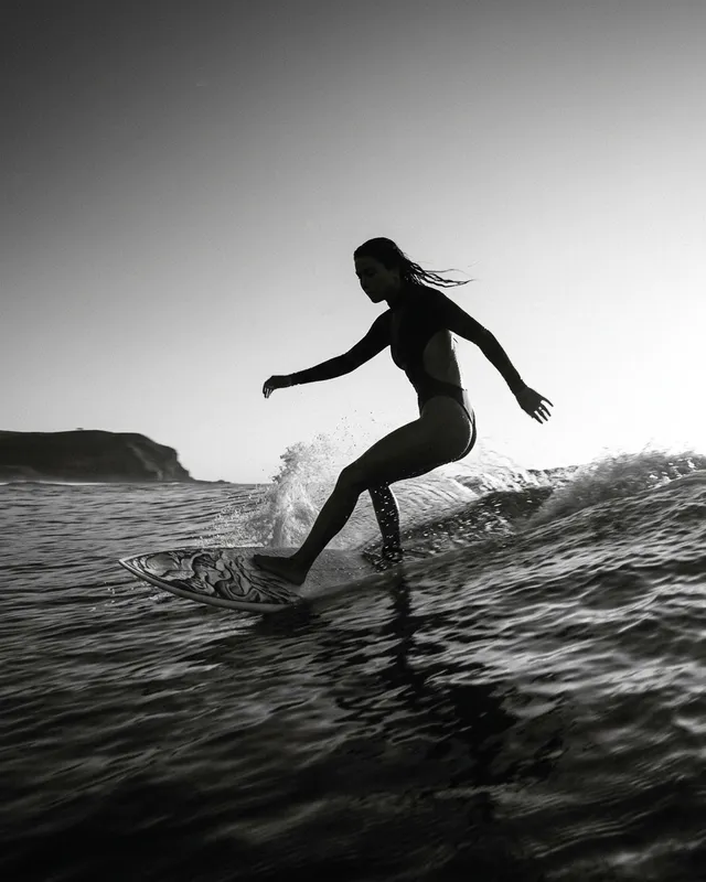 A dynamic black-and-white editorial photograph captures a silhouette of a surfer riding a wave at sunset, evoking a sense of freedom and adventure. The composition highlights the fluid movement and the contrast between the dark ocean and the soft light on the horizon.