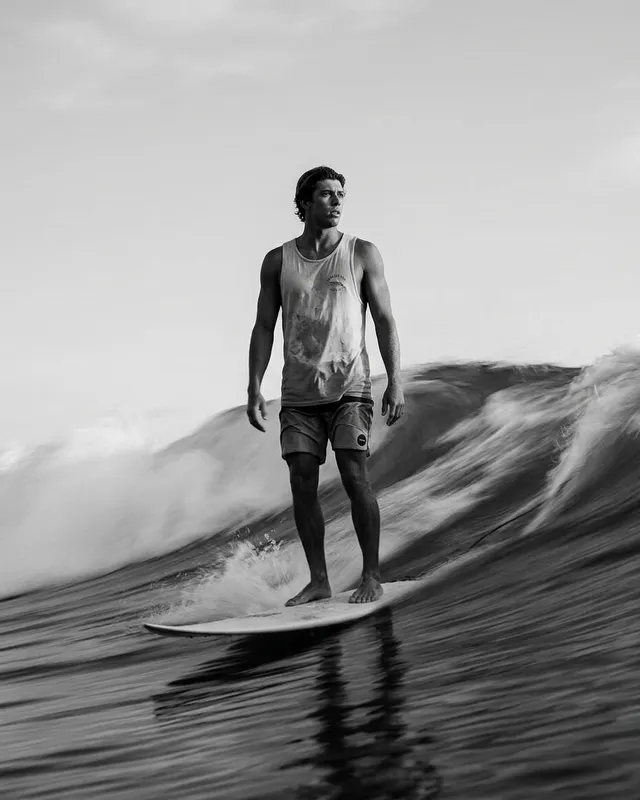 A dynamic black and white photograph capturing a male surfer skillfully riding a wave, conveying a sense of movement and adventure. The image features a moody atmosphere with shades of gray and contrasting shadows.