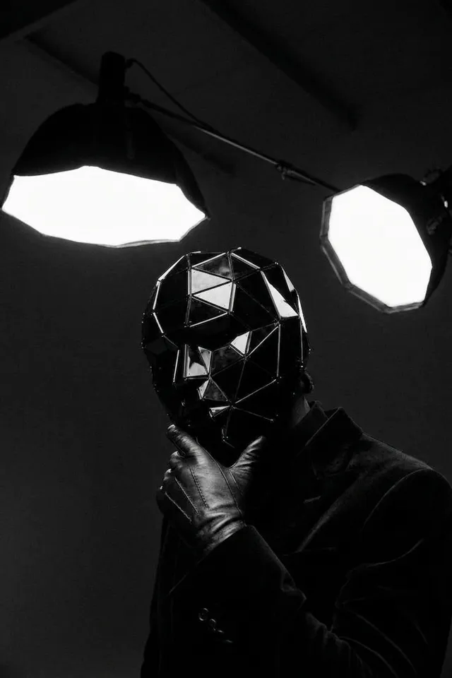 A mysterious figure in a geometric black mask poses thoughtfully under bright spotlights, captured in a striking black and white editorial style. The dominant colors are deep blacks and stark whites, creating an enigmatic and dramatic mood.