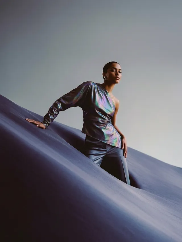 A high-fashion editorial photograph features a model striking a pose atop a dramatic, undulating background. The image is characterized by iridescent hues and deep blues, creating a sleek and sophisticated mood.