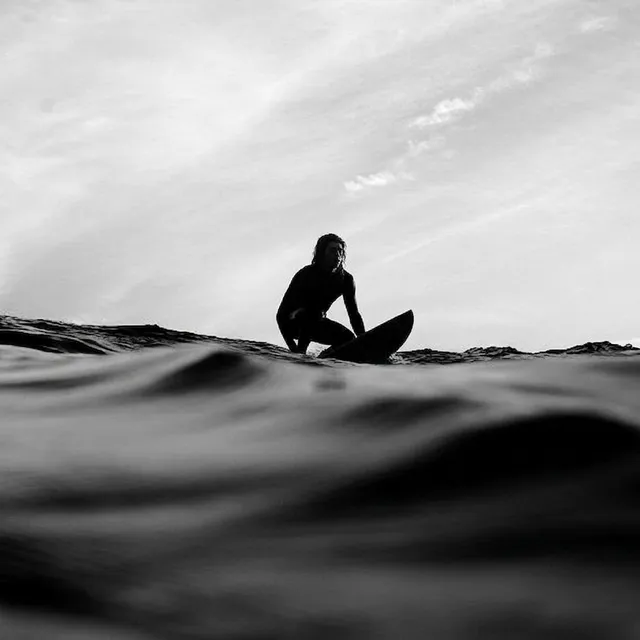 A striking black-and-white editorial photograph captures a lone surfer poised on their board against the backdrop of rolling waves, conveying a sense of serenity and solitude. The dominant shades of gray and the dramatic sky amplify the mood of calm introspection.