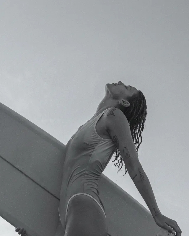 A black and white photograph capturing a woman with a surfboard, evoking a sense of strength and serenity against a minimalistic background. The mood leans toward the raw and adventurous.