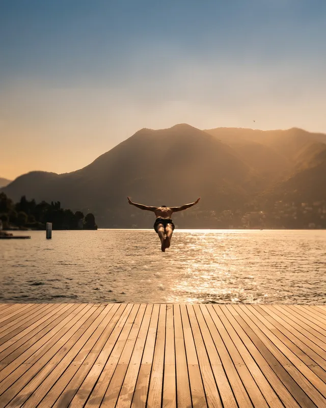 A silhouette of a person diving off a wooden dock into a tranquil lake, with majestic mountains in the background. The scene is illuminated by warm golden hues, creating a serene and adventurous mood.