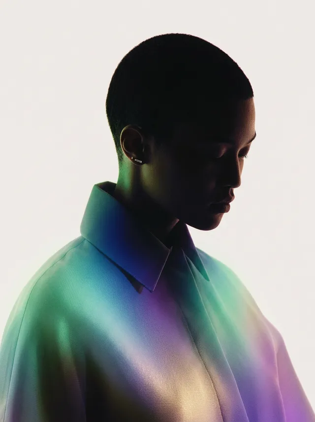 This editorial photography piece features a silhouette of a figure wearing an iridescent shirt, set against a soft, white background. The mood is contemplative, with a palette of pastel blues, purples, and greens creating a serene and modern aesthetic.