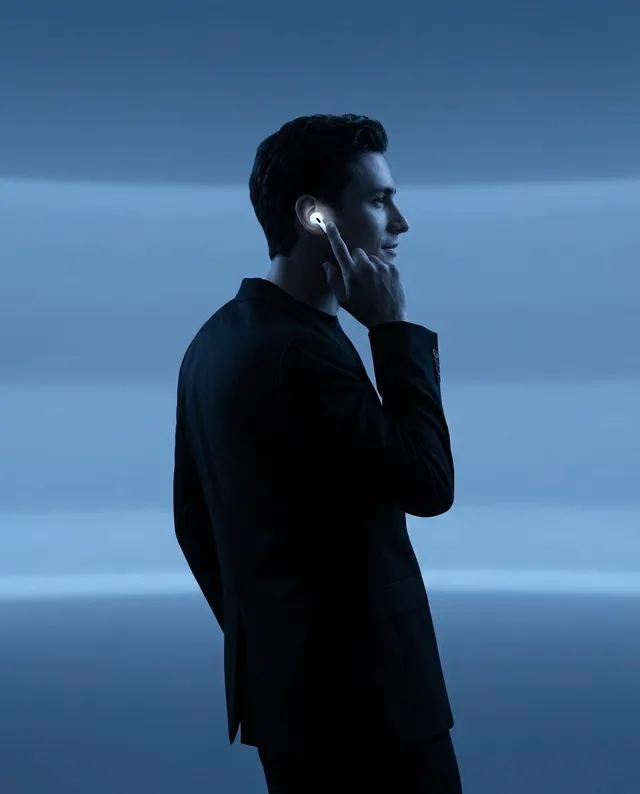 A moody portrait in studio photography featuring a man with a sleek hairstyle, wearing a suit, and interacting with wireless earbuds against a backdrop of soft blue lighting.