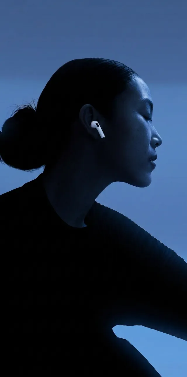 A minimalist editorial photograph features a profile of a woman with a sleek bun, wearing wireless earbuds against a deep blue backdrop. The mood is serene and modern, highlighting the contrast between the soft lighting and dark silhouette.