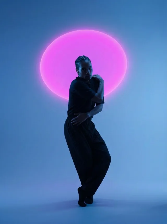 A striking editorial photograph features a dancer silhouetted against a vibrant pink circular light, set against a cool blue backdrop. The mood is ethereal and dynamic, highlighted by the contrast of the soft pink and deep blue hues.
