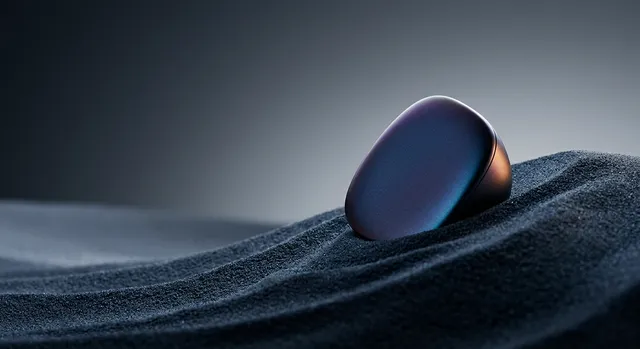 A sleek, metallic object rests on a textured surface of dark sand, captured in a modern, high-contrast editorial photography style. The mood is minimalistic and sophisticated, with dominant shades of deep blue, black, and subtle highlights of iridescent color.