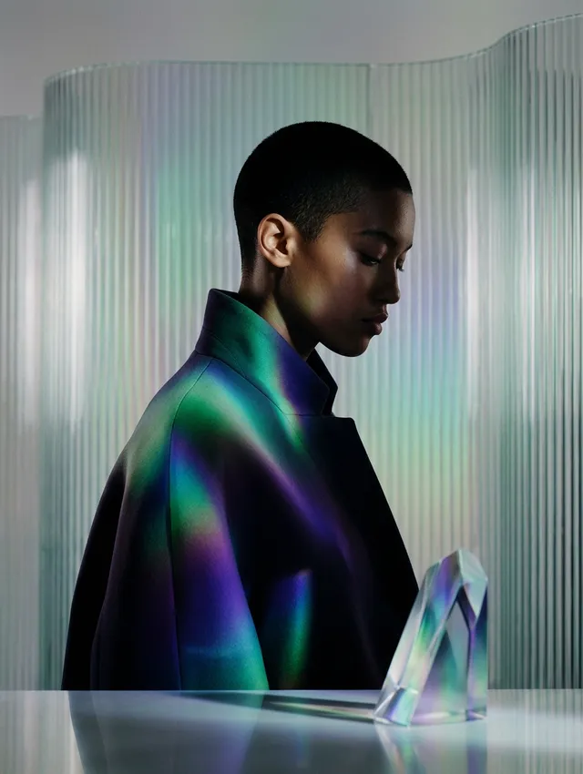 A striking editorial photograph features a profile of a model with a shaved head, dressed in a dark coat, illuminated by iridescent light that casts vibrant colors across her face and surroundings. The mood is introspective and futuristic, with dominant hues of deep blue, green, and purple.