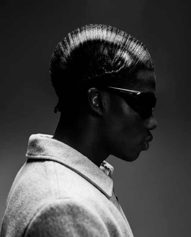 A striking black-and-white editorial portrait captures the profile of a stylish individual with a sleek hairstyle and sunglasses, exuding a mood of sophistication and coolness. The dominant tones of deep black and subtle gray enhance the dramatic, minimalist aesthetic.