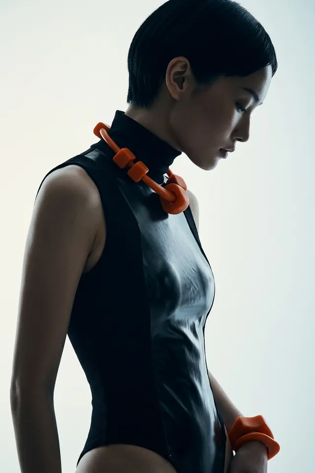 This editorial photograph features a side profile of a model sporting a glossy black bodysuit, complemented by bold orange accessories. The soft lighting creates a modern, minimalist mood, emphasizing the sleek lines and textures.