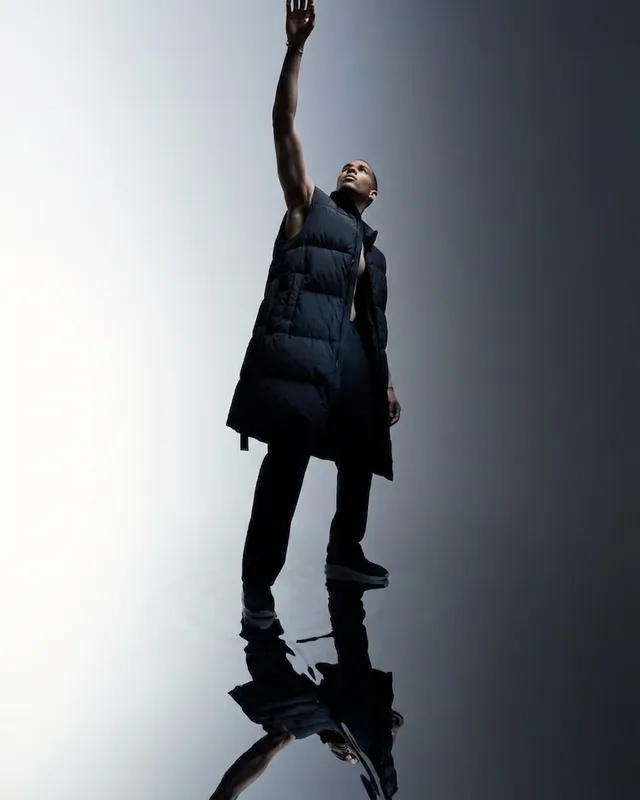 A striking editorial photograph features a model in a sleeveless black puffer coat, reaching upward against a stark, reflective background that enhances the dramatic mood. The dominant colors are deep blacks and soft grays, creating a sleek and modern aesthetic.