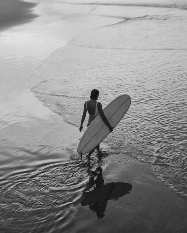 A monochromatic photograph capturing a woman carrying a surfboard while wading through the water at the shore. The scene conveys a serene mood with muted tones of gray and soft reflections.