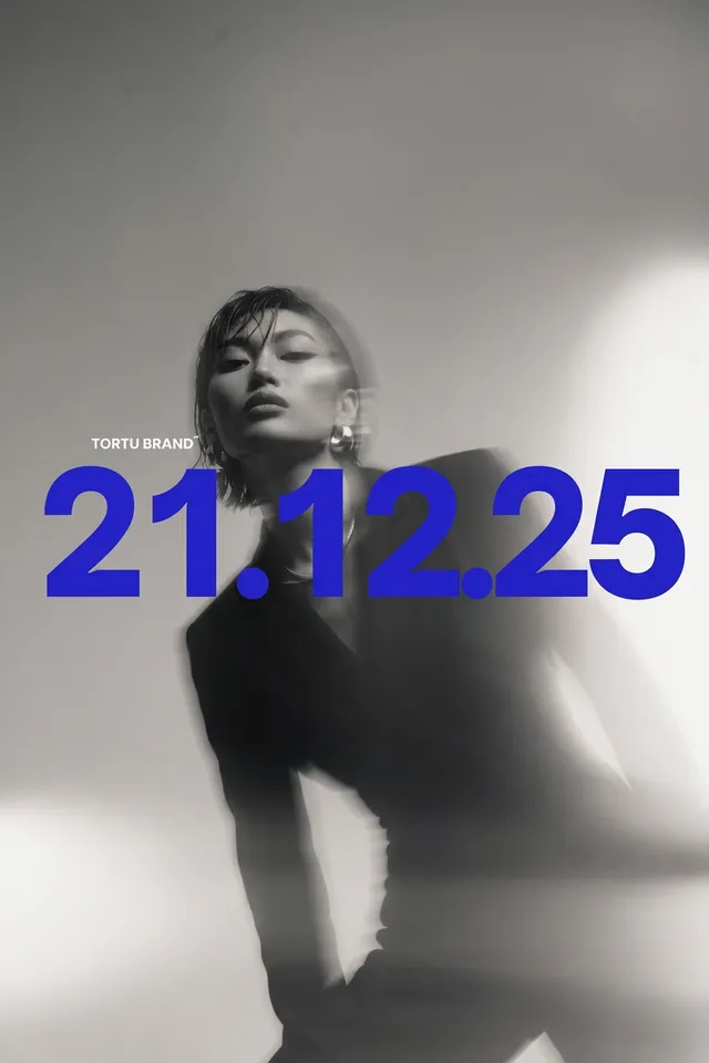 A sleek, editorial photograph featuring a model in a black suit, captured in soft focus to evoke a sense of movement and intrigue. The dominant colors are monochromatic shades of black and white, contrasted by bold blue text displaying "21.12.25," creating a modern and stylish mood.