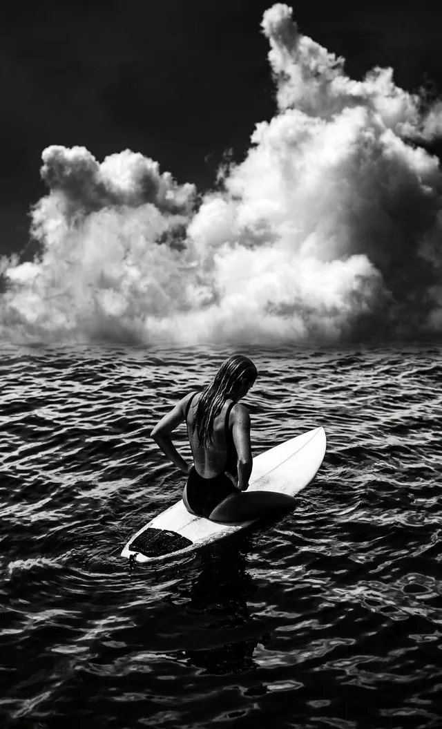 A monochromatic editorial photograph features a lone surfer sitting on a surfboard amidst dark, turbulent waters, with dramatic clouds swirling overhead. The mood is introspective and moody, highlighted by contrasting shades of black and white.