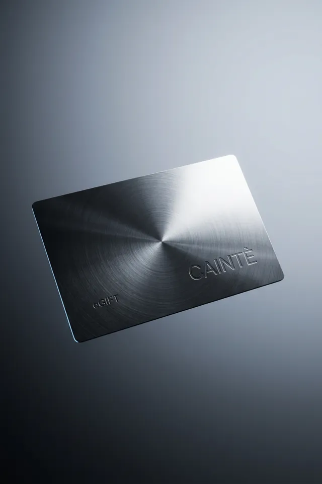 A sleek, metallic gift card features a minimalist design with the word "CAINTE" embossed on its surface. The image employs a high-contrast, editorial photography style with cool gray tones, conveying a modern and sophisticated mood.